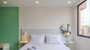 Ein Hotelzimmer mit Bett, Nachttisch mit Lampe, Spiegel und Blick auf die Stadt durch ein Fenster.