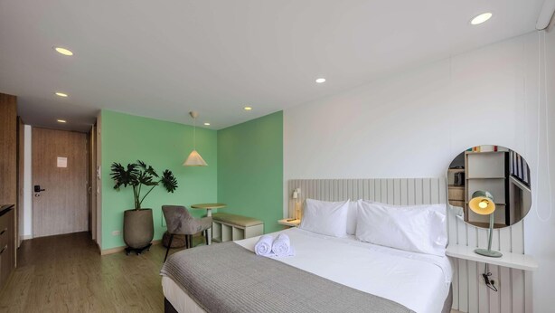 Ein Hotelzimmer mit Bett, Nachttisch, Lampe, Blumentopf, Stuhl und einer Tür zu einem anderen Raum.