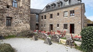 A stone building with a gravel courtyard, red chairs, and a stack of firewood. - Oberburg | Unique vacation apartment in a historic castle. (Kobern)