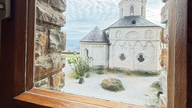 A stone building with a tower and a smaller structure in front, viewed through a wooden-framed window. - Oberburg | Unique vacation apartment in a historic castle. (Kobern)