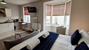 Ein Hotelzimmer mit Bett, einem kleinen Tisch, einem Stuhl, einer Küchenzeile und einem Fenster mit Jalousien.