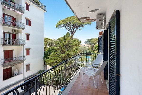 Ein Balkon mit Metallgeländer, einem weißen Stuhl und Blick auf Bäume und Gebäude.