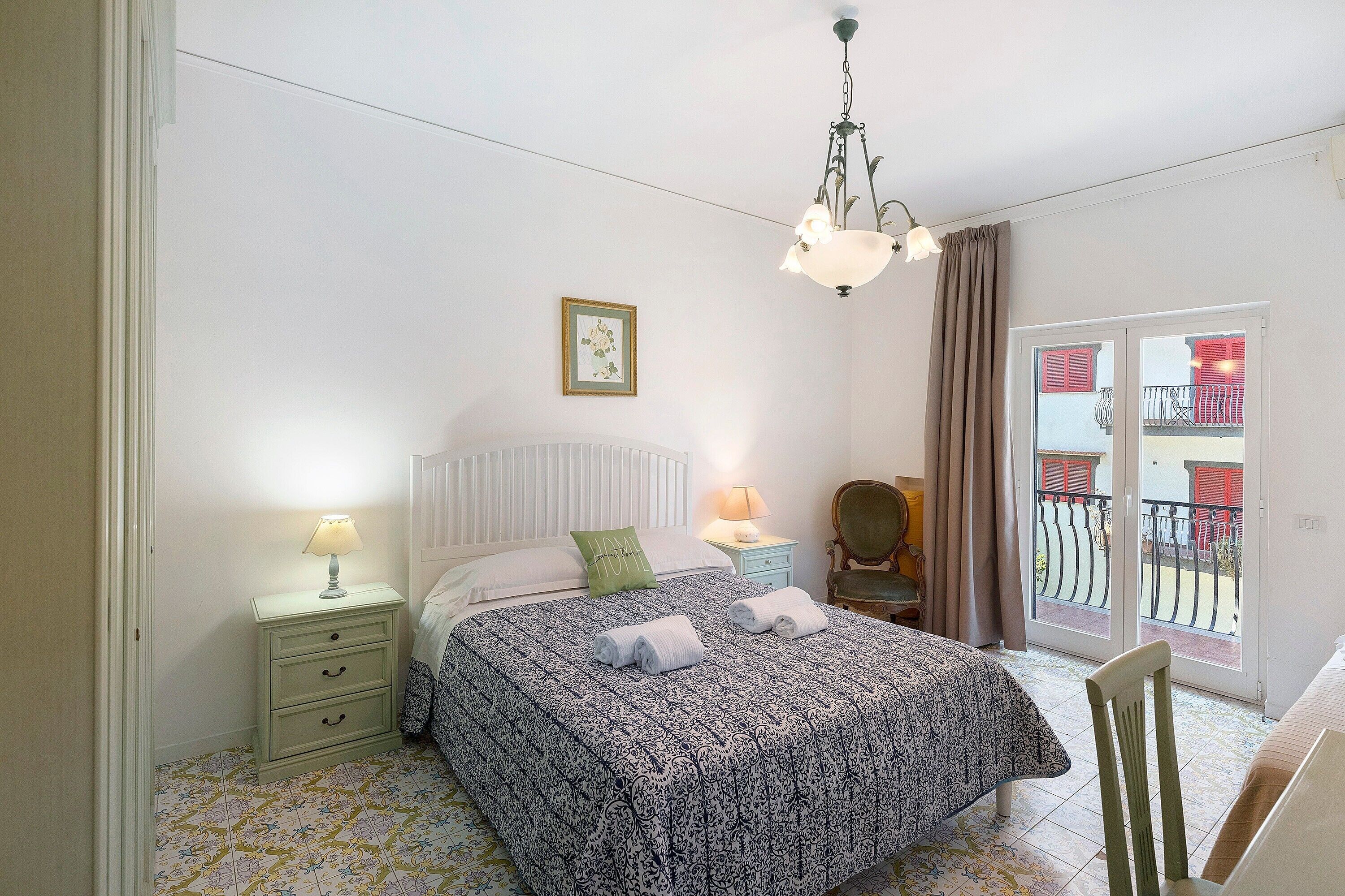 Un dormitorio con cama, mesitas de noche, una silla, vistas al balcón y una lámpara colgante.