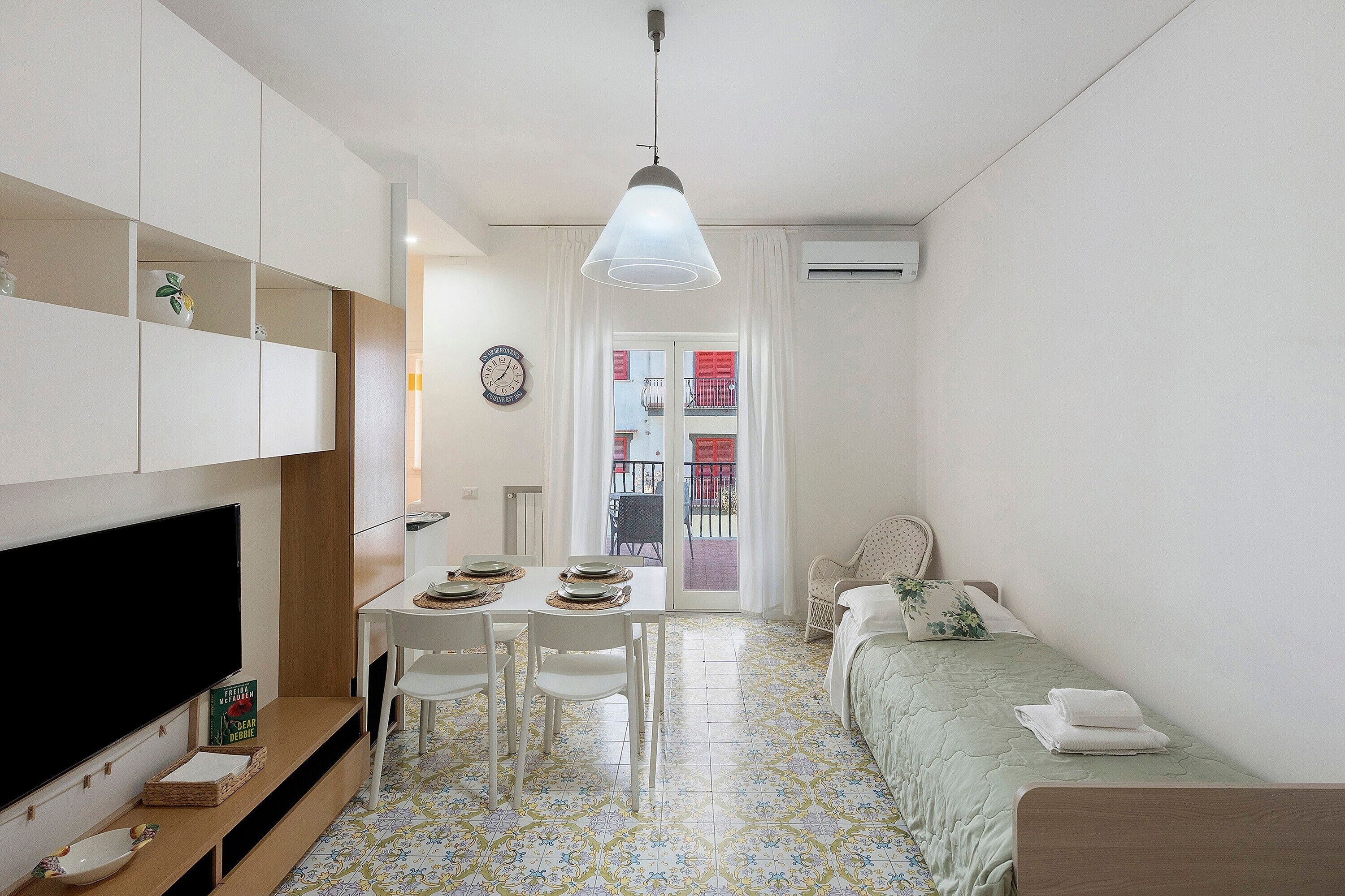 Quarto compacto com televisão, mesa de jantar, cadeiras, uma cama e vista para o exterior.