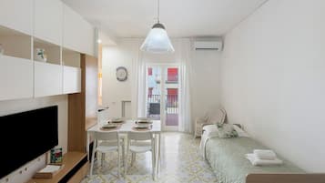 Quarto compacto com televisão, mesa de jantar, cadeiras, uma cama e vista para o exterior.