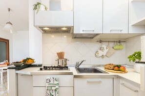 Moderni keittiö, jossa on valkoiset kaapit, ruostumattomasta teräksestä valmistettu pesuallas, kaasuliesi ja seinälle ripustettu kattoteline.