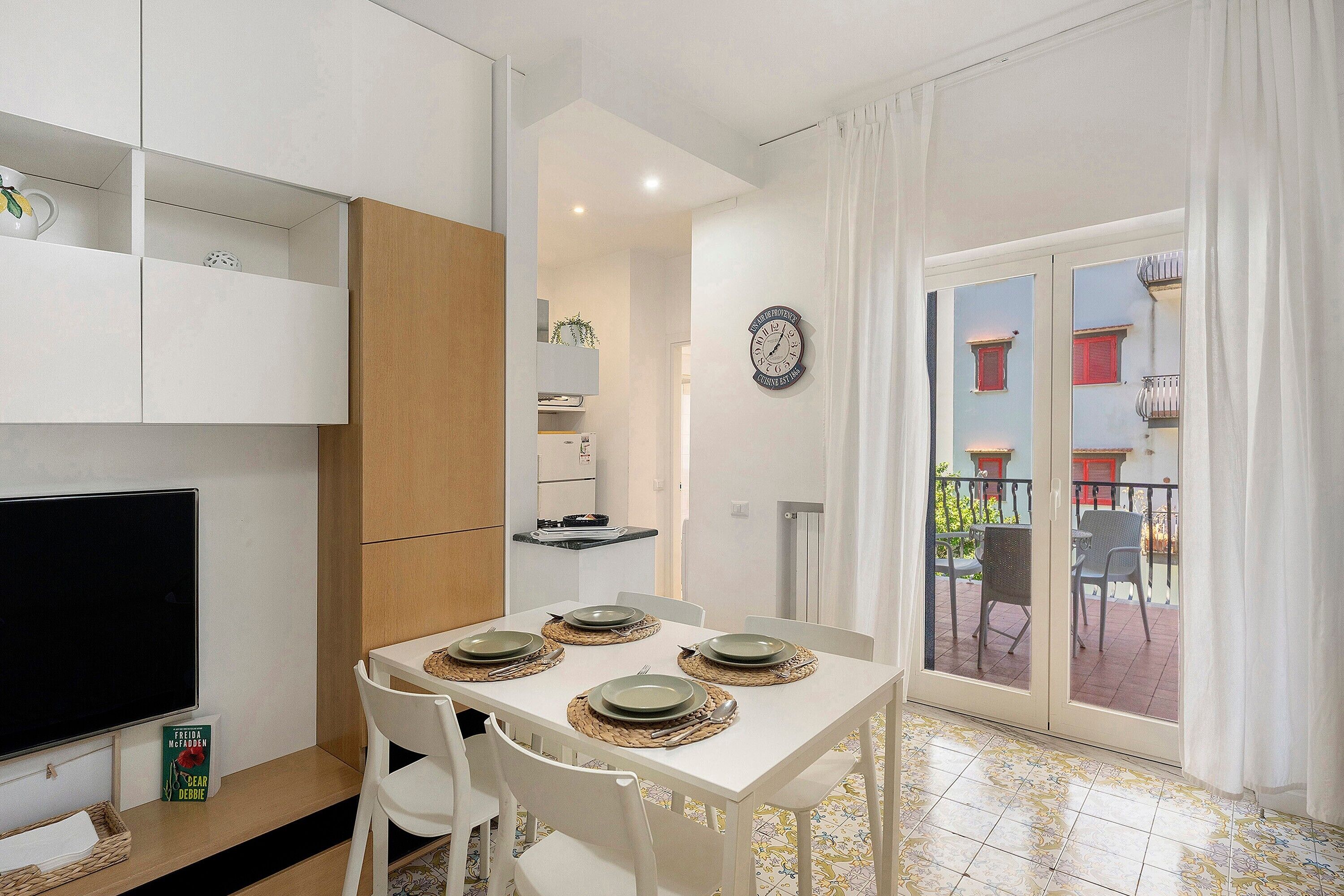 A dining area with a table set for four, white chairs, a television, and a kitchen area with cabinets and a clock on the wall.