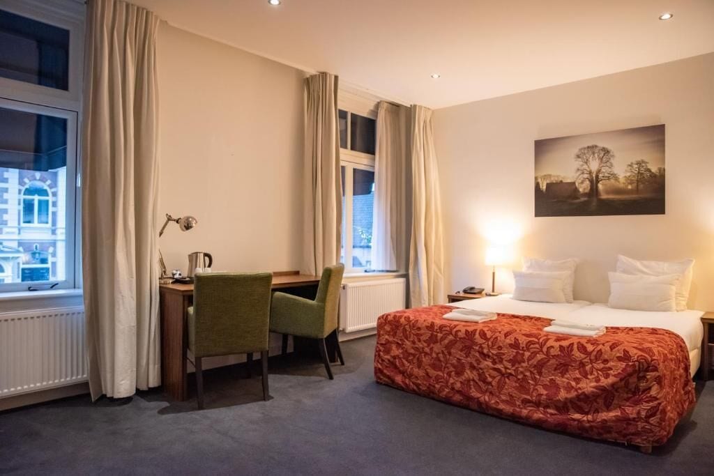 A hotel room with a bed, a desk, a chair, a window with curtains, and a painting on the wall.