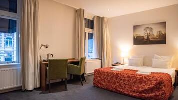 A hotel room with a bed, a desk, a chair, a window with curtains, and a painting on the wall.