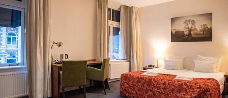 Habitación de hotel con cama, escritorio, silla, ventana con cortinas y un cuadro en la pared.