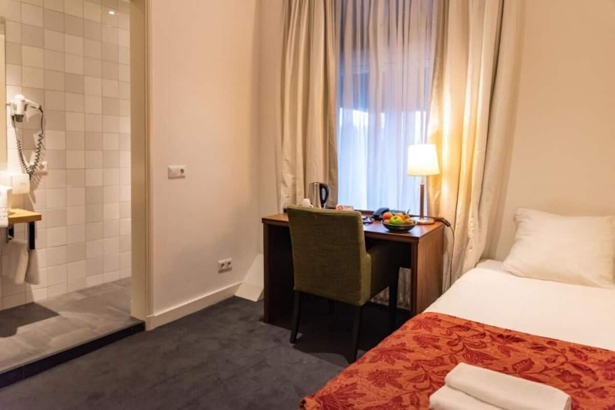 Ein Hotelzimmer mit Bett, Schreibtisch mit Stuhl, einem Fenster mit Vorhängen, einer Lampe und einer Dusche.