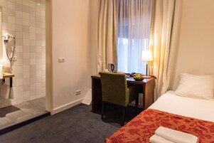 Habitación de hotel con cama, escritorio con silla, ventana con cortinas, lámpara y zona de ducha.
