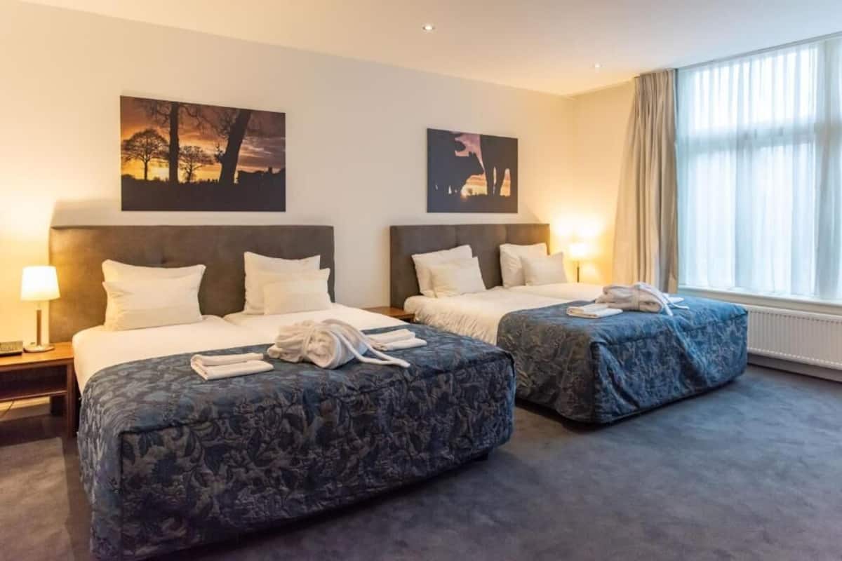 Ein Hotelzimmer mit zwei Betten, einem großen Fenster mit Vorhängen und Bildern an der Wand.