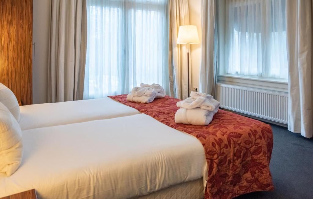 Una habitación de hotel con dos camas, una colcha roja, una lámpara y una ventana con cortinas.