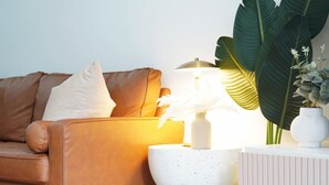 Un canapé en cuir marron avec un coussin blanc, une table d’appoint blanche à motifs et une lampe, ainsi qu’un vase blanc contenant une plante.