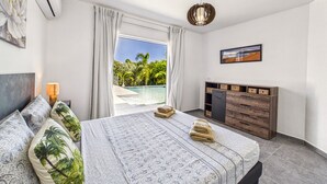 Un dormitorio con cama, mesita de noche, lámpara, cómoda de madera, cuadro y vistas a una piscina y follaje.