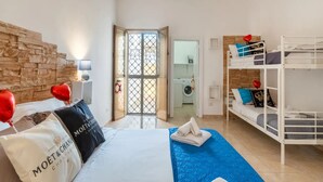 A bedroom with a bed, a blue quilt, a white headboard, a wooden wall, a white ceiling light, a white door, and a laundry area visible through the door. - Brezza Salentina House Giardino Privato (Porto Cesareo)