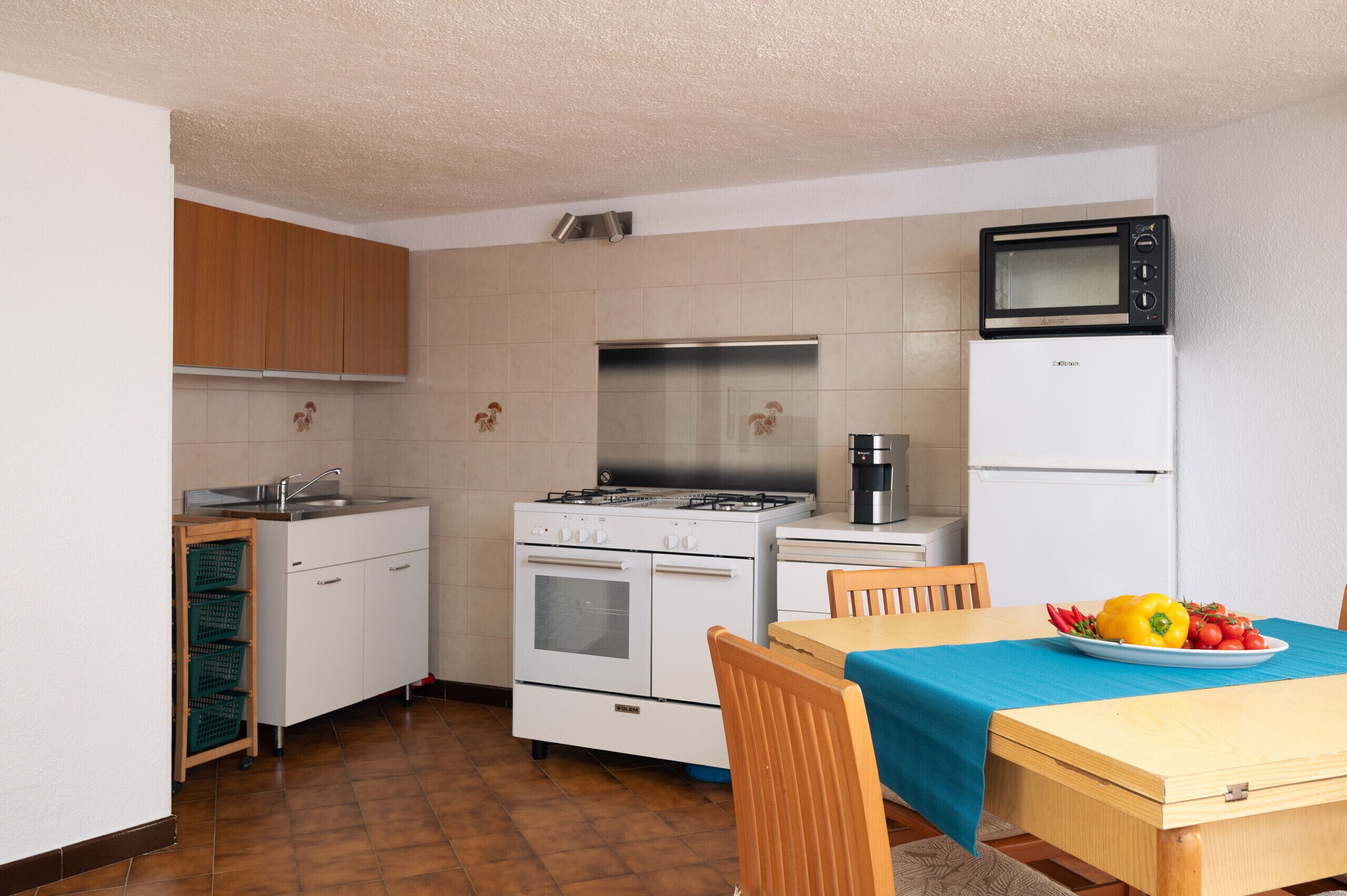 A compact kitchen with white appliances, a microwave, and a stove. There is a dining table with a blue tablecloth and a plate of fruit.
