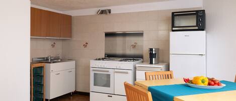 A compact kitchen with white appliances, a microwave, and a stove. There is a dining table with a blue tablecloth and a plate of fruit.