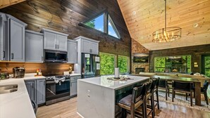 A modern kitchen with grey cabinets, a central island, and a dining area with wooden chairs. - New! 5BR cabin w/Hot Tub*Theater*Arcade*Fireplace (Pigeon Forge)