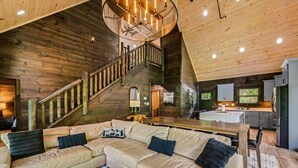 A log cabin interior with a wooden staircase, a sectional sofa, a kitchen area, and a dining table. - New! 5BR cabin w/Hot Tub*Theater*Arcade*Fireplace (Pigeon Forge)