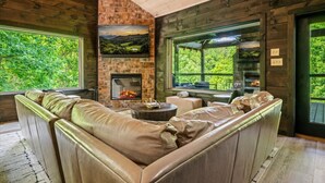 A cozy room with a stone fireplace, leather sofa, and large windows offering a view of greenery. - New! 5BR cabin w/Hot Tub*Theater*Arcade*Fireplace (Pigeon Forge)