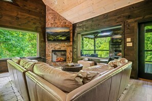 Ein gemütliches Zimmer mit Kamin aus Stein, Ledersofa und großen Fenstern mit Blick ins Grüne.