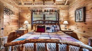 Ein Schlafzimmer in einer Blockhütte mit einem Bett, zwei Nachttischen, einem Fenster mit Jalousien und einem Holz-Kopfteil.