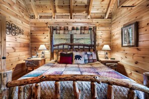 Een slaapkamer in een blokhut met een bed, twee nachtkastjes, een raam met jaloezieën en een houten hoofdbord.