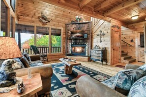 Een knus cabin interieur met een open haard, houten wanden, een trap en een groot raam met uitzicht op groen.