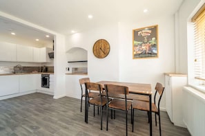 A modern kitchen with white cabinets, a wooden dining table, and four chairs. A large wall clock and a movie poster are also visible.