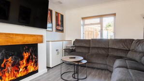 Un salon avec un canapé gris, une petite table ronde, un téléviseur fixé au mur et une cheminée où brûle un feu.