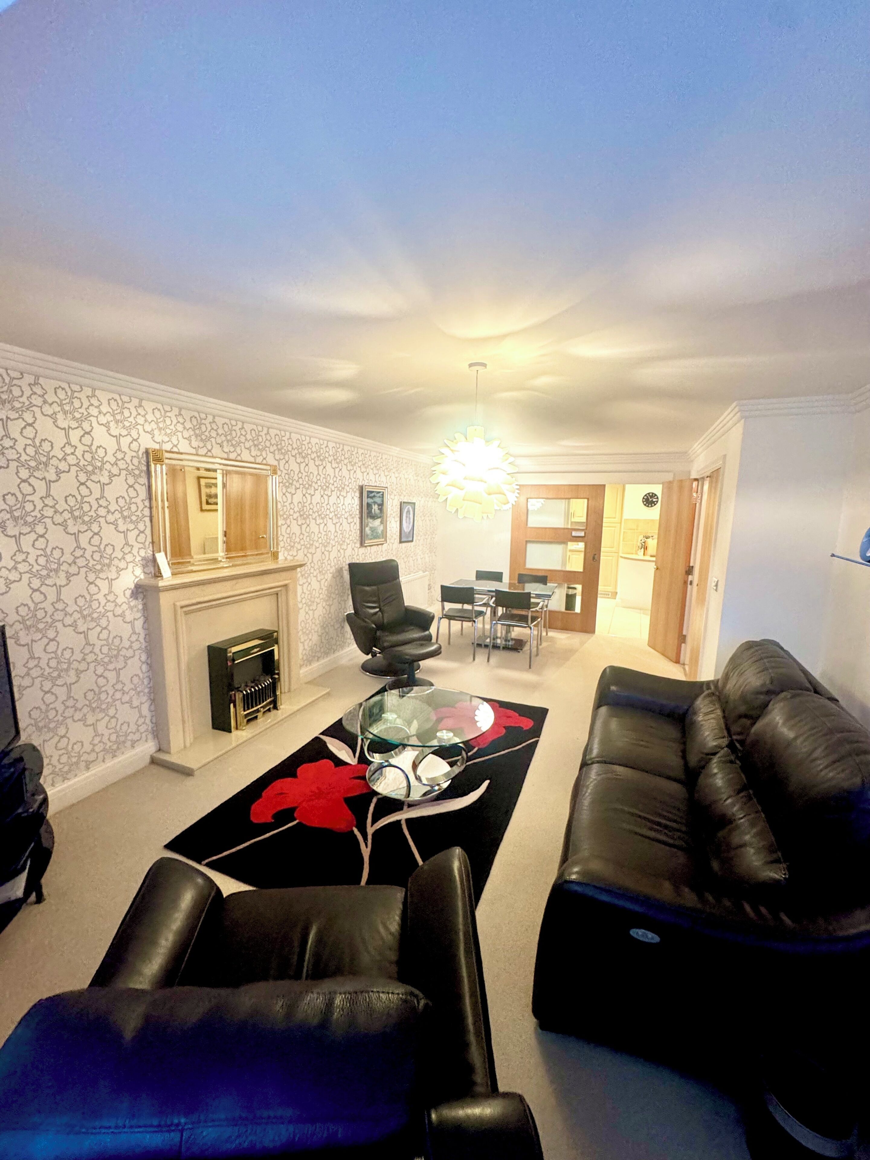 A living room with a fireplace, a glass coffee table, a black leather sofa, and a dining area in the background.