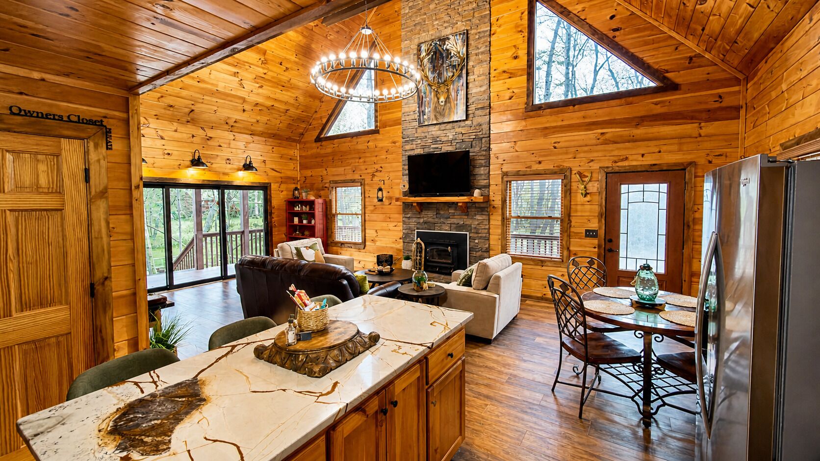 A cabin interior with a kitchen, dining area, and living room, featuring wooden walls, a stone fireplace, and large windows.