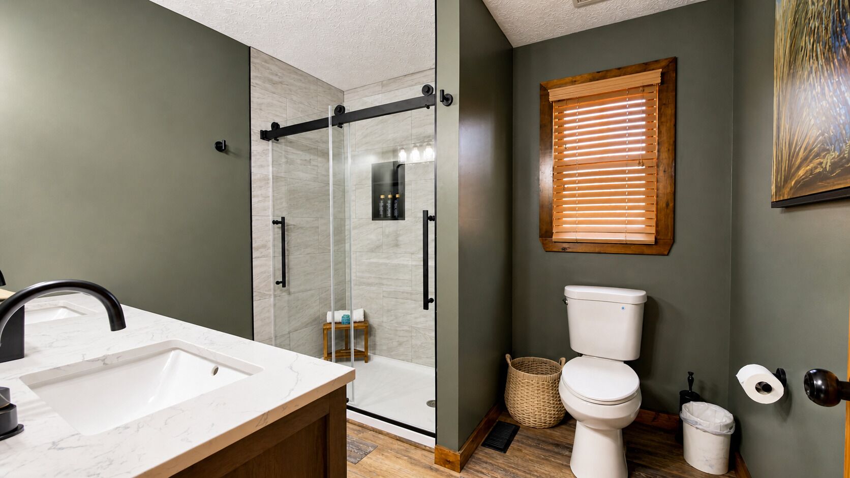 A bathroom with a glass-enclosed shower, a sink, a toilet, and a window with blinds.