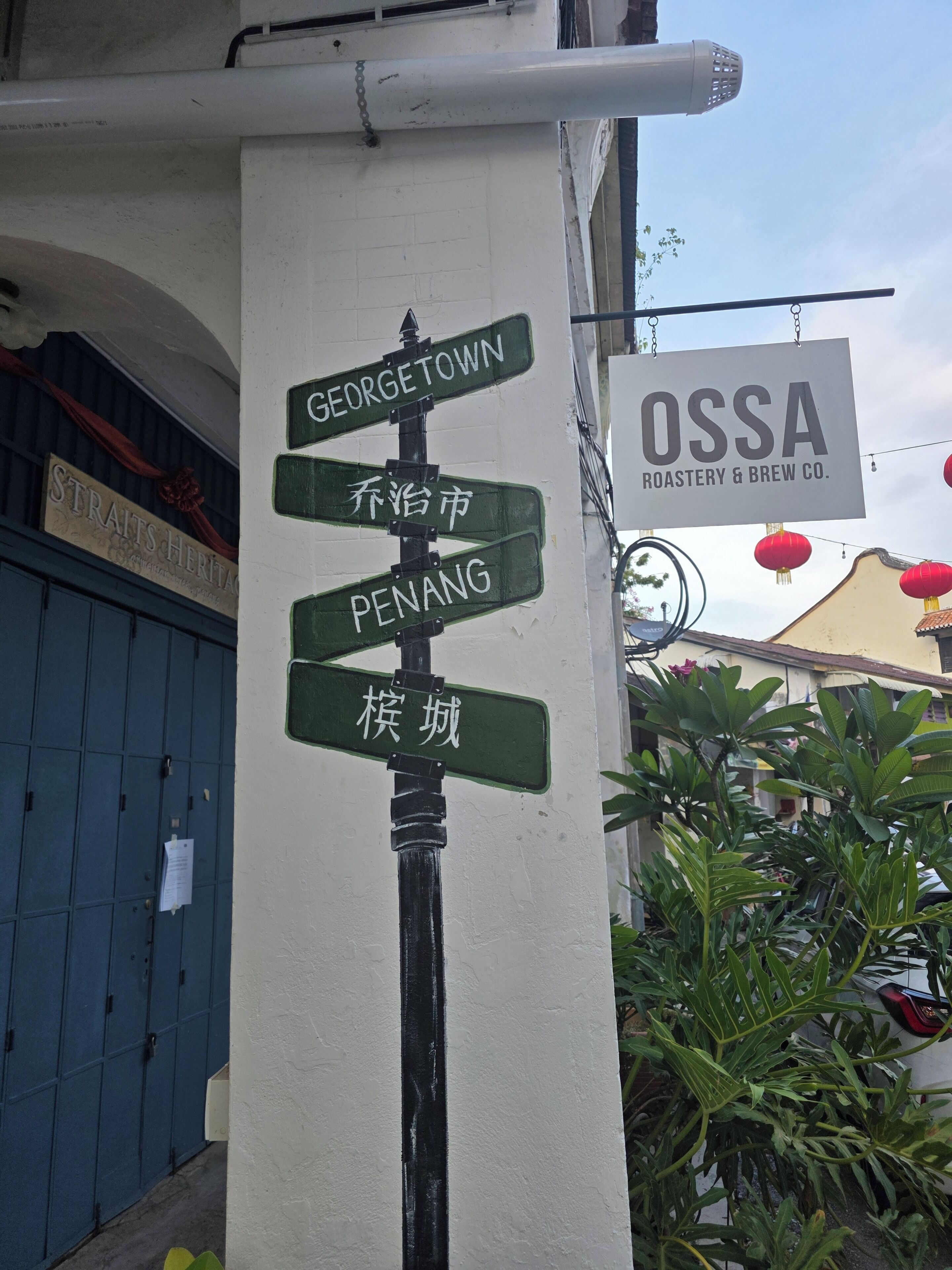 Un panneau indicateur indiquant plusieurs destinations, un bâtiment situé à proximité avec une enseigne pour OSSA Roastery & Brew Co., et une porte bleue en arrière-plan.