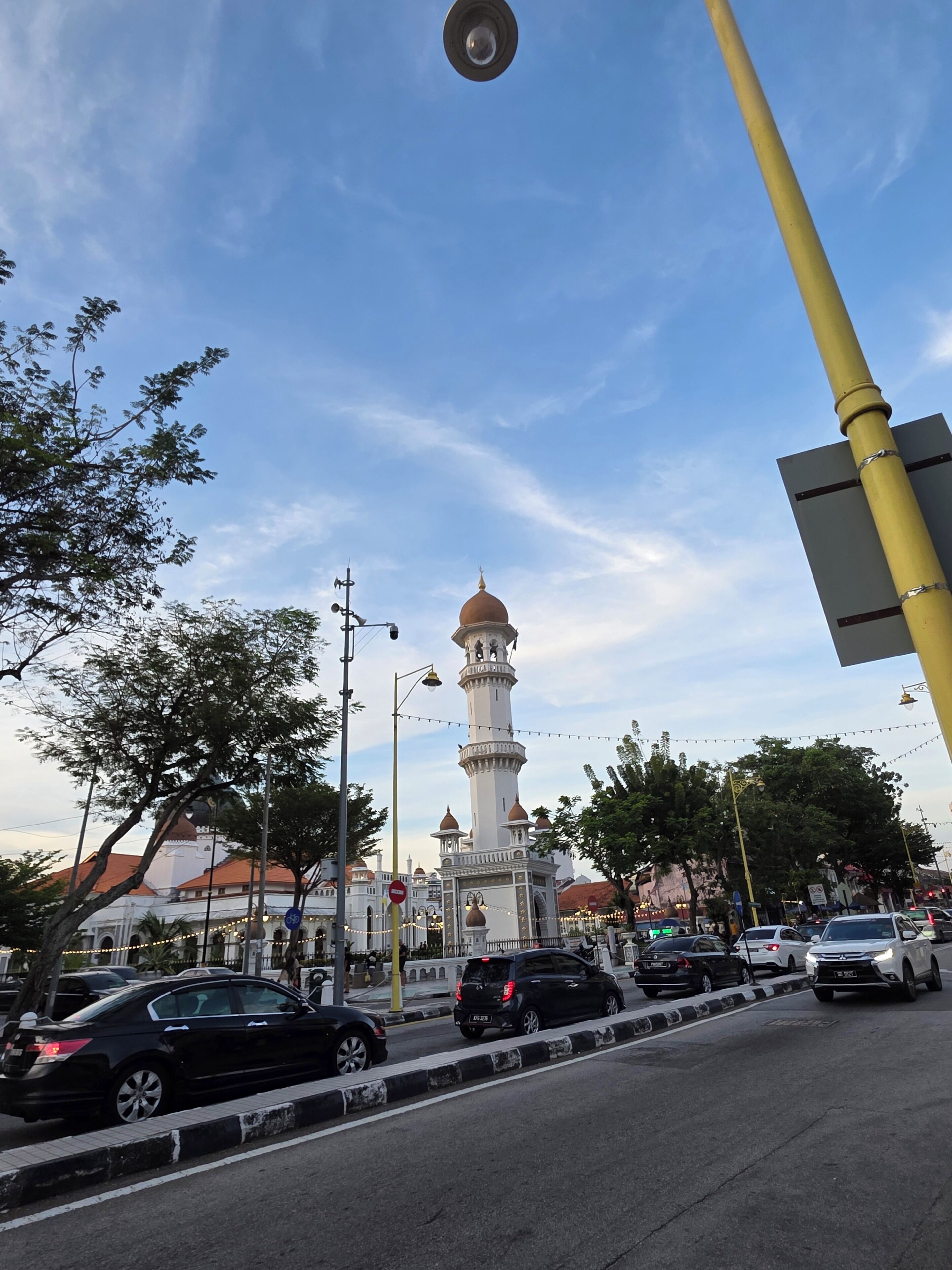 A street view with a prominent white tower with a dome and minarets, surrounded by trees and streetlights.