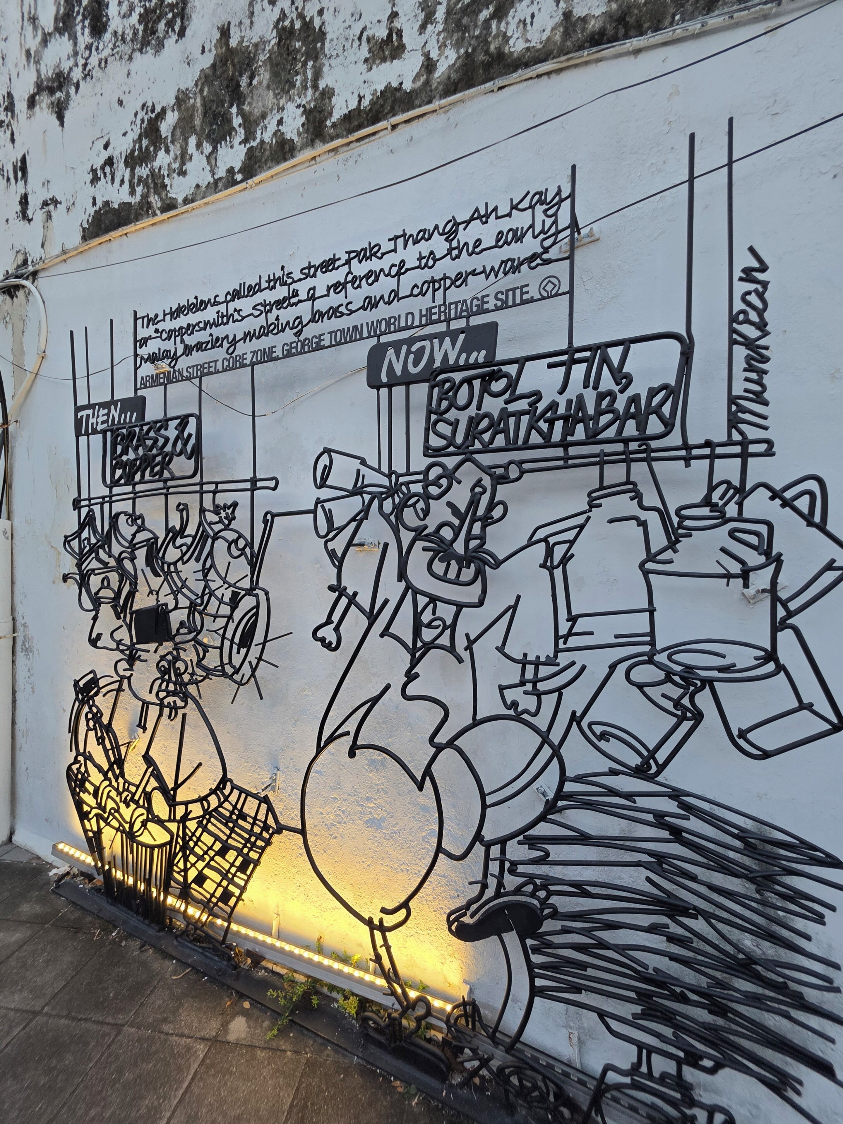 A wall with a historical mural depicting brass and copper workers, including a sign that reads 'Now... BOTOL, SURATKABAR.'