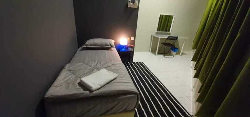A single bed with a pillow and a folded blanket, a bedside table with a lamp, a small desk with a chair, and a window with curtains.