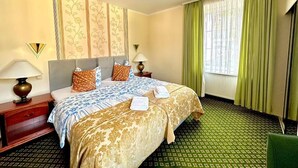 Une chambre d’hôtel avec un lit, des tables de chevet, des lampes, une fenêtre avec des rideaux et un tapis à motifs.