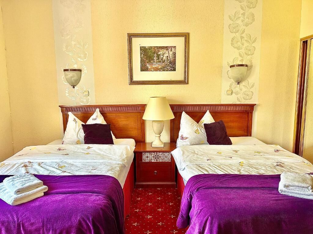 Ein Hotelzimmer mit zwei Betten, einem Nachttisch, einer Lampe, einem gerahmten Bild an der Wand und einem roten Teppich.