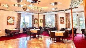 Une salle à manger d’hôtel avec de nombreuses tables et chaises, de grandes fenêtres avec des rideaux et une moquette rouge.