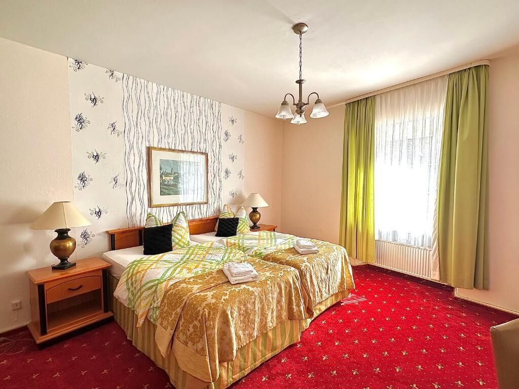 A hotel room with a double bed, two bedside tables with lamps, a red carpet, a window with curtains, and a wall with a patterned wallpaper.