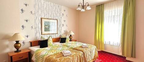 A hotel room with a double bed, two bedside tables with lamps, a red carpet, a window with curtains, and a wall with a patterned wallpaper.