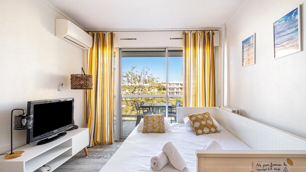 A hotel room with a bed, a TV, a balcony with yellow curtains, and a sign on the wall. - Air-conditioned studio with sea view and pool (Antibes)