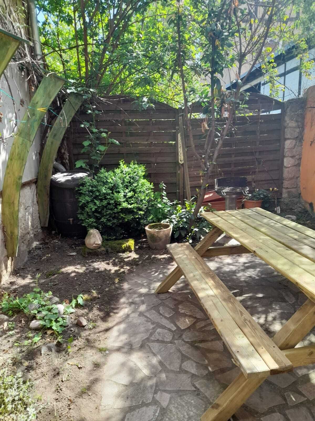 Ein Holztisch für ein Picknick in einem Garten mit einer mit Steinfliesen ausgelegten Terrasse, umgeben von Topfpflanzen und einem Holzzaun.