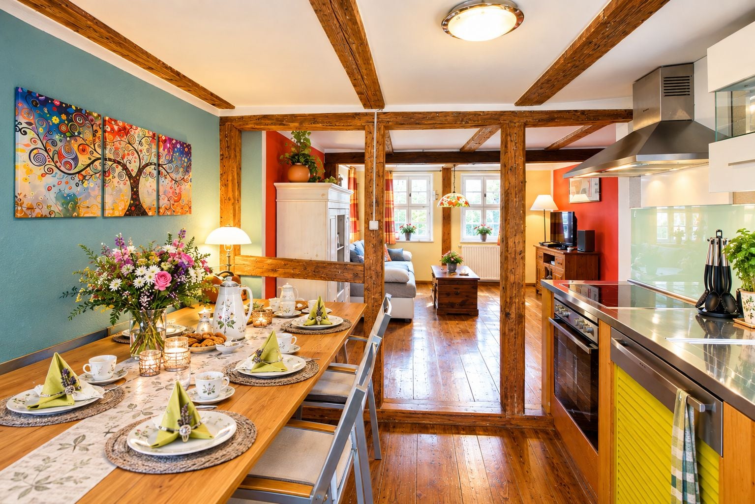 Ein Essbereich mit Holztisch, gedeckt für eine Mahlzeit, eine Küche mit modernen Geräten und ein Wohnzimmer mit Sofa und einem Bild an der Wand.