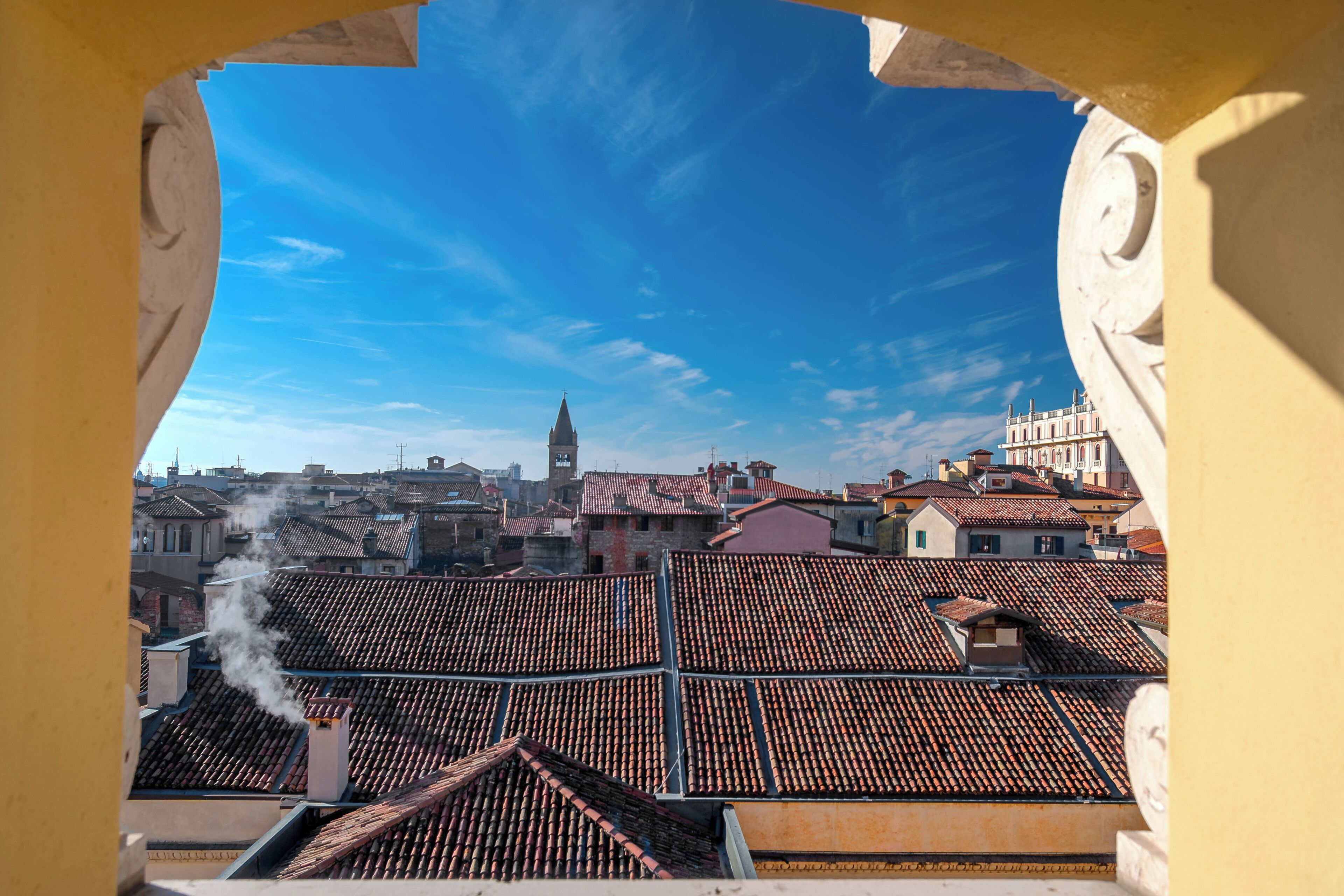 A view through an archway of a cityscape with tiled rooftops, a prominent tower, and a clear sky.