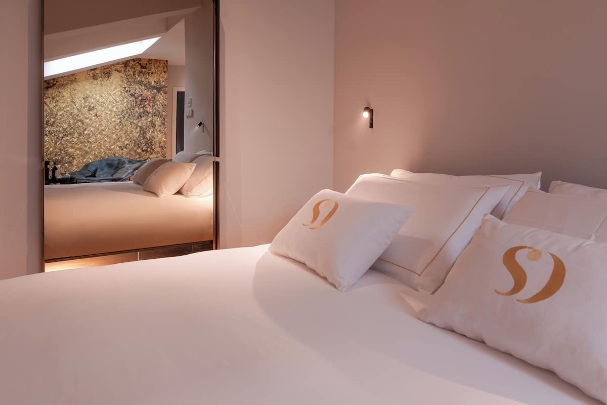 A modern hotel room with a large bed, a wall-mounted light, and a view of another room through an open door.