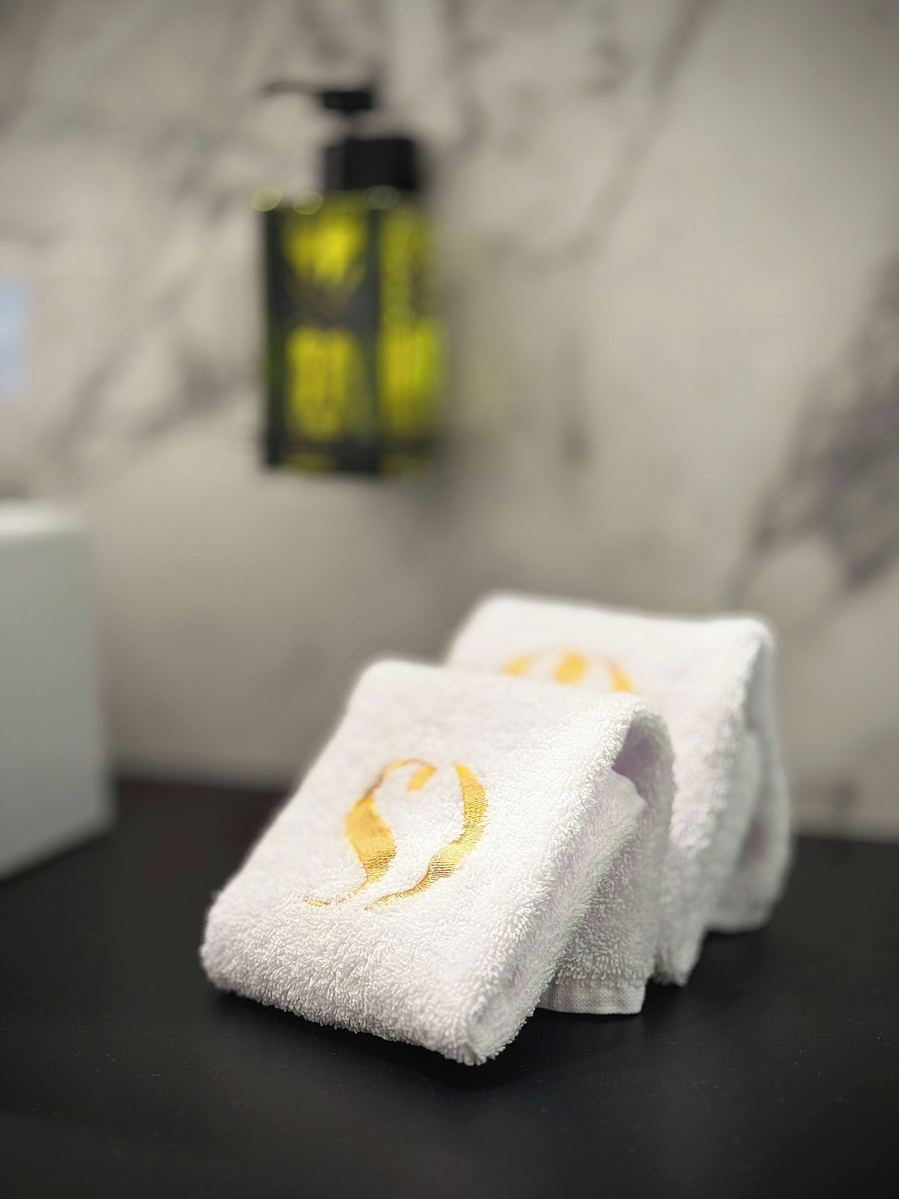 Two white towels with a golden emblem on a dark surface, a bottle of liquid soap, and a marble wall in the background.